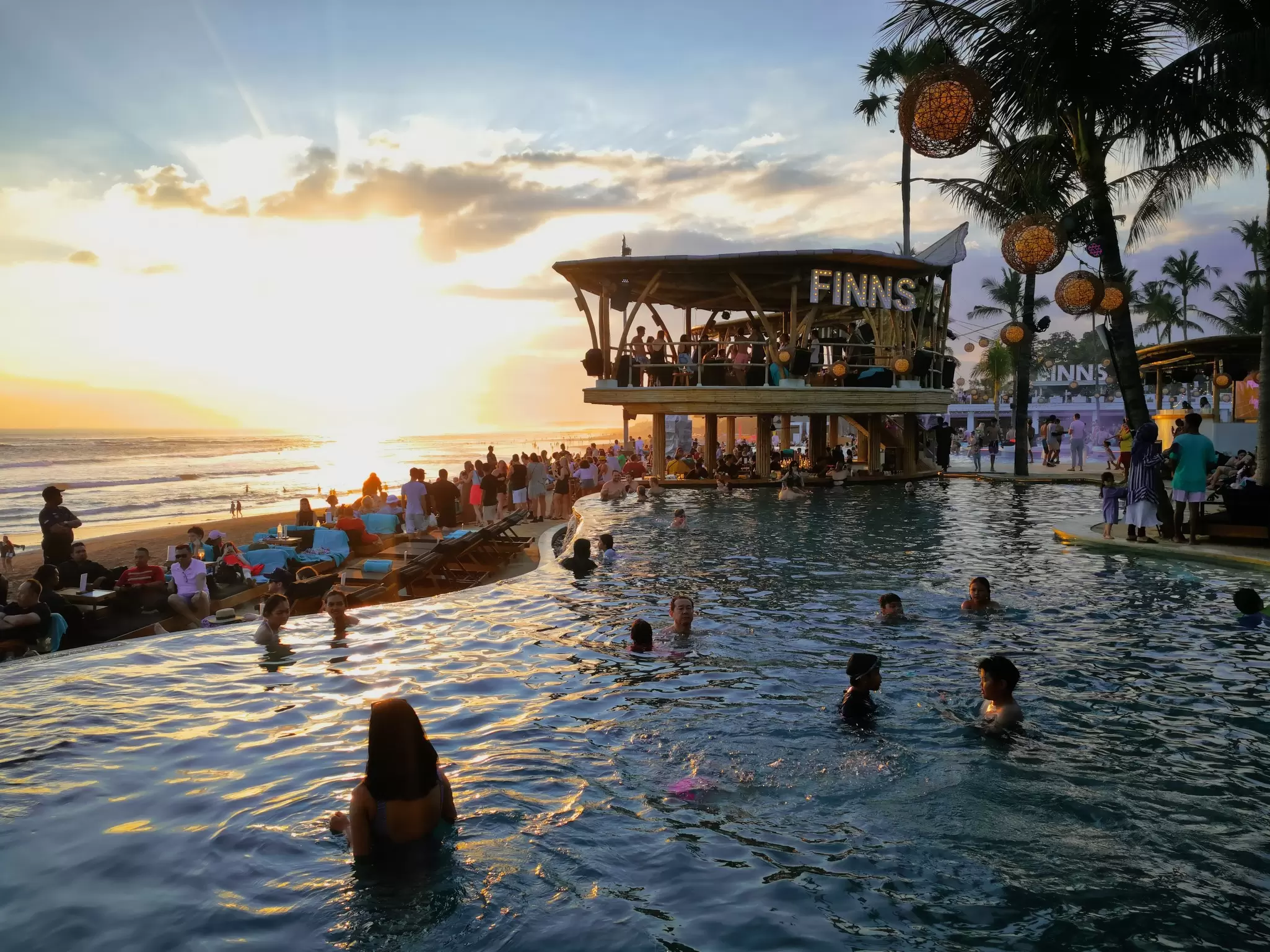 People standing in a large pool amid palm trees near a beach. People stand on a two-tier structure nearby that appears to be made of bamboo with the word "Finns" affixed to the top tier