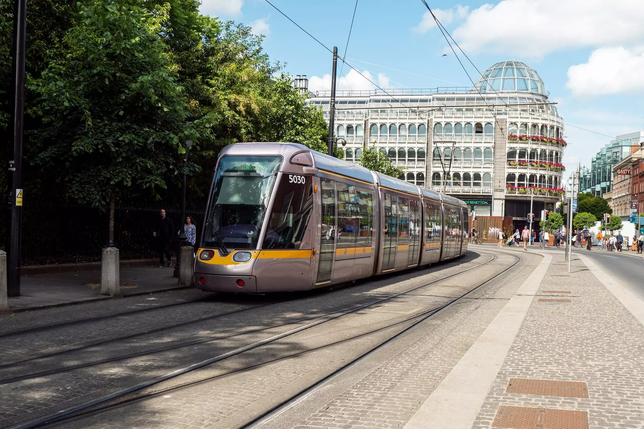 A light rail train passes through a city center. People walk around in the background in front of a building with many windows.