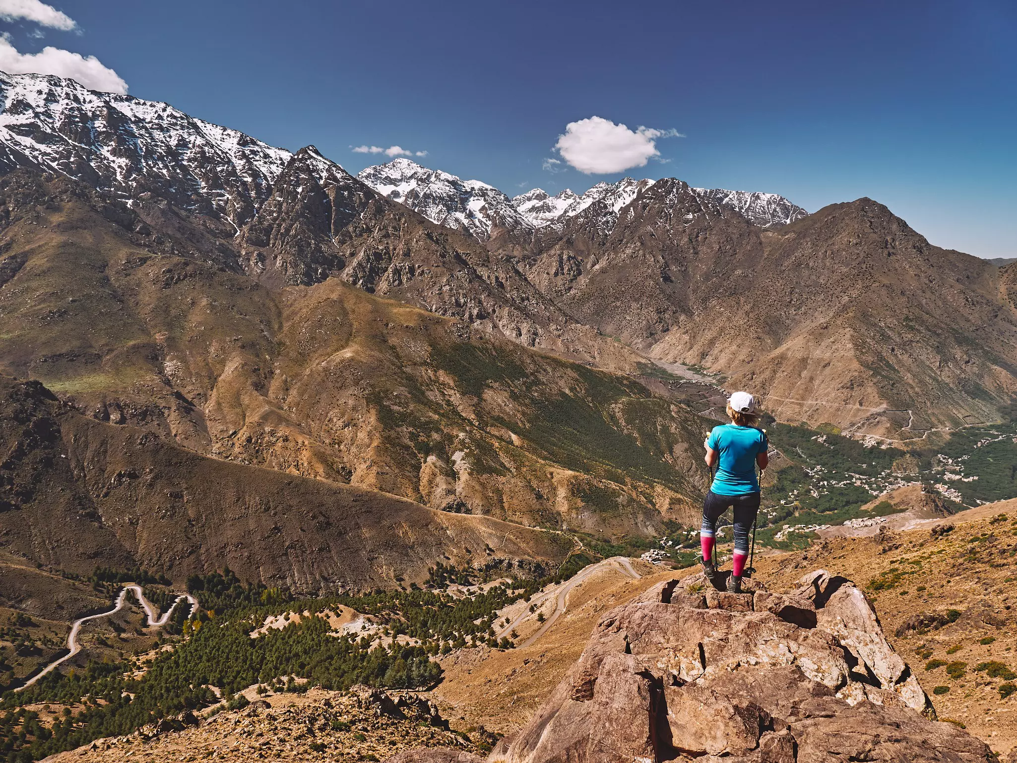 Caucasian blonde tourist girl on a hike looking towards snow covered High Atlas monuntain peaks with Jebel Toubkal in the middle in Morocco Africa