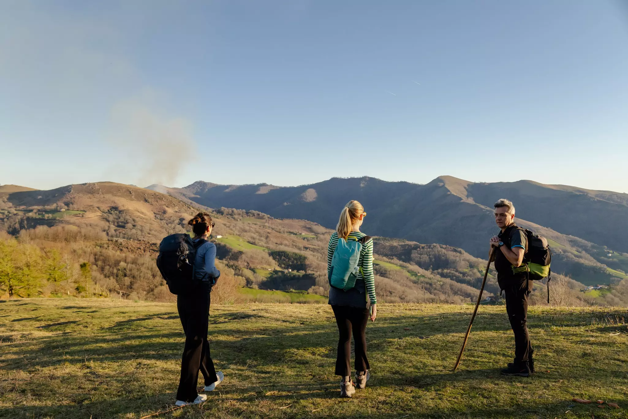 Three people walk on green grass in a mountainous landscape; one holds a walking stick.