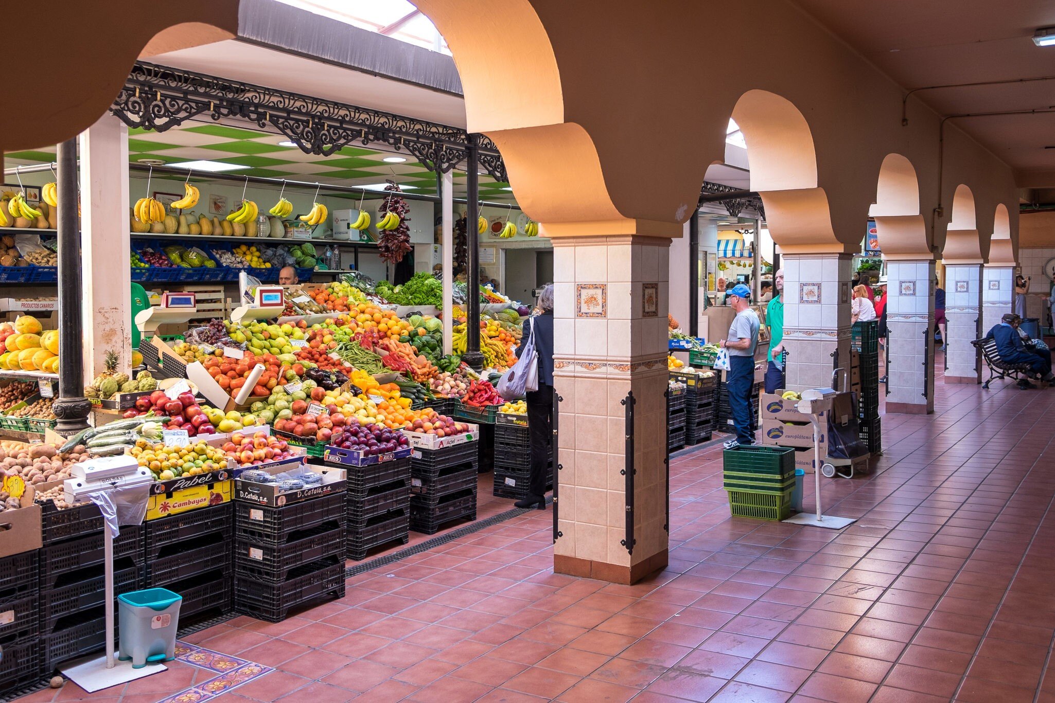 Stall piled with bananas, apples and citrus beside a tiled walkway lined with salmon-coloured arches and shoppers