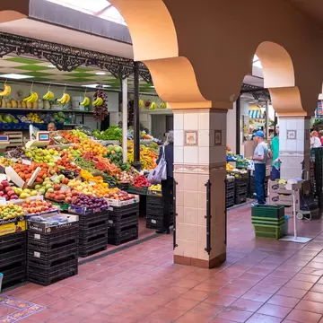 Stall piled with bananas, apples and citrus beside a tiled walkway lined with salmon-coloured arches and shoppers