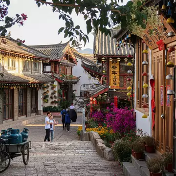 Stone-paved lane between tiled wooden houses with hanging bells, pink flowers and a few people walking in late-day sun.