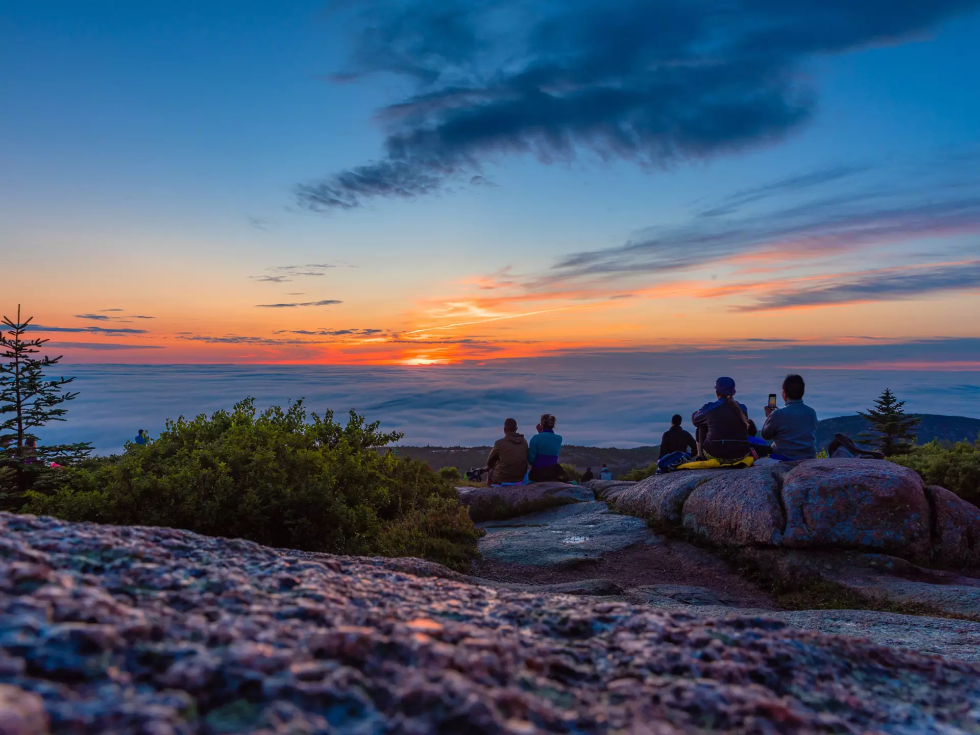 People in silhouette sit at a mountain viewpoint looking out at the sky as the sun rises above the clouds casting an orange glow everywhere; Cadillac Mountain, Acadia National Park, Maine.