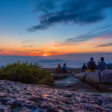  People in silhouette sit at a mountain viewpoint looking out at the sky as the sun rises above the clouds casting an orange glow everywhere; Cadillac Mountain, Acadia National Park, Maine.