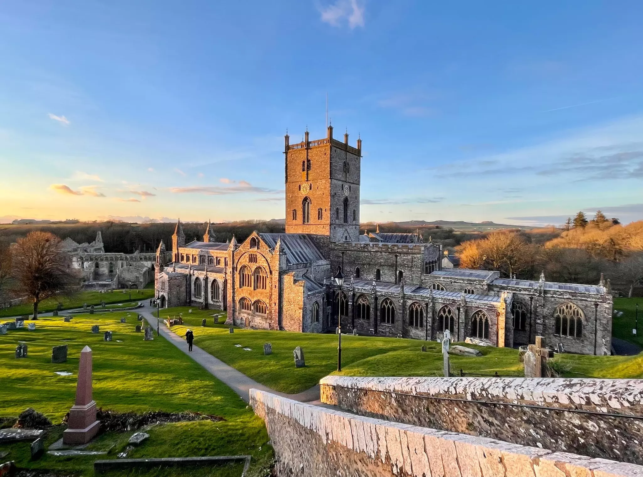 Wide shot of stone cathedral and palace with a concrete walkway in the foreground and wide-spaced graves in green grass as the sun rises or sets.