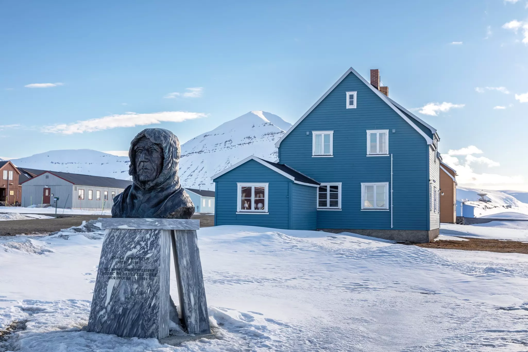 A bronze bust of a man stands on a plinth in a village. Snow covers the ground, and snowy mountains are pictured in the distance.