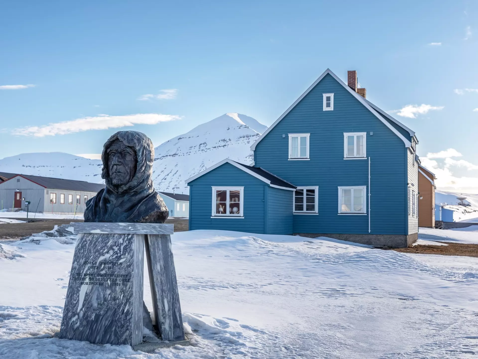 A bronze bust of a man stands on a plinth in a village. Snow covers the ground, and snowy mountains are pictured in the distance.