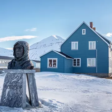 A bronze bust of a man stands on a plinth in a village. Snow covers the ground, and snowy mountains are pictured in the distance.