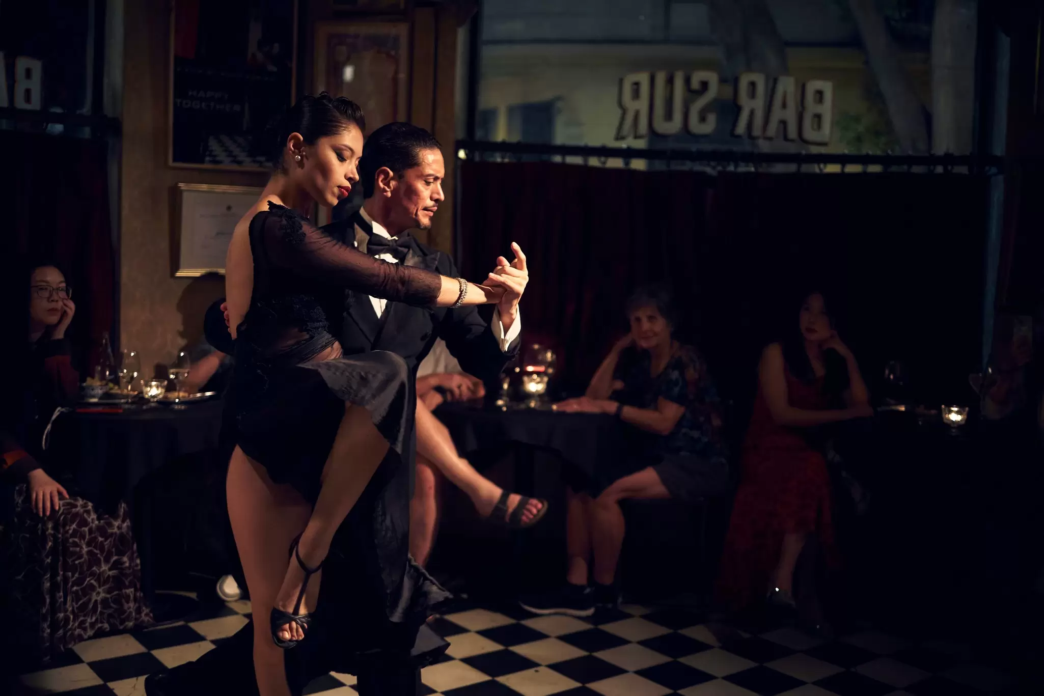 Tango dancers perform in a venue with people watching them from tables in the background.