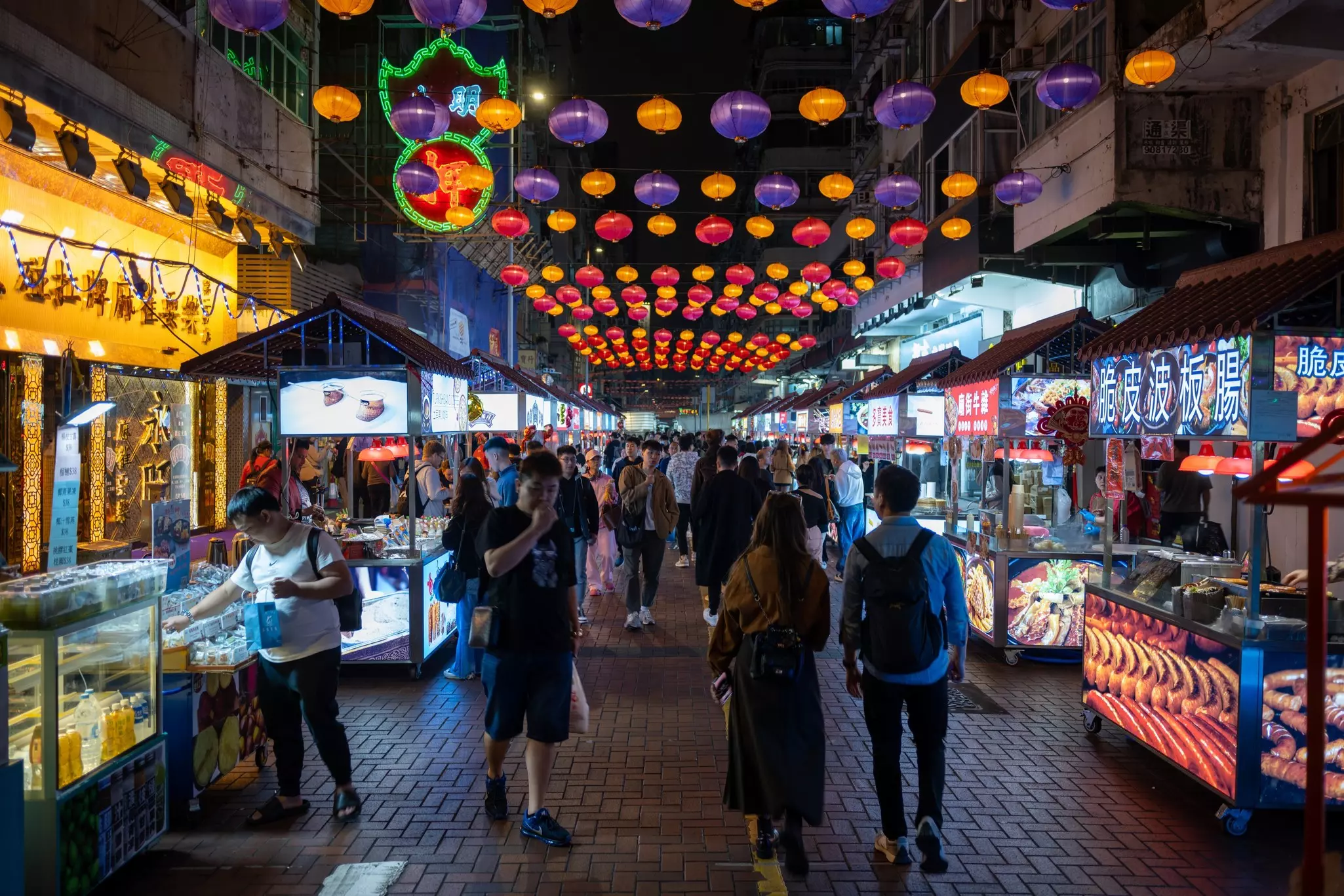 People walk past food stands at a night market, with multicolored lights overhead.