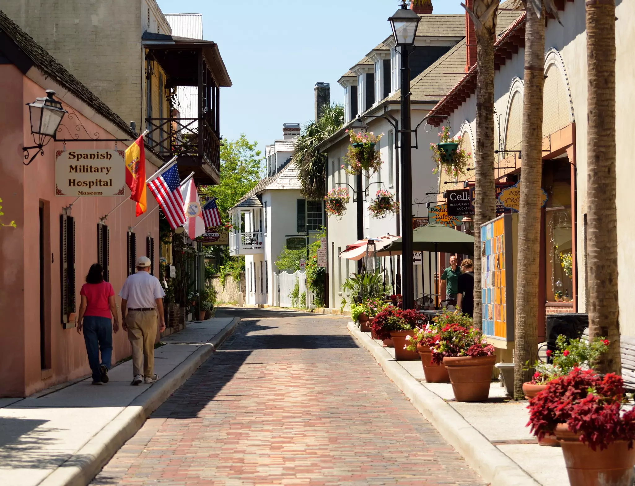 Two people wander down a quaint historic street where flags fly from a museum with the sign "Spanish Military Hospital"
