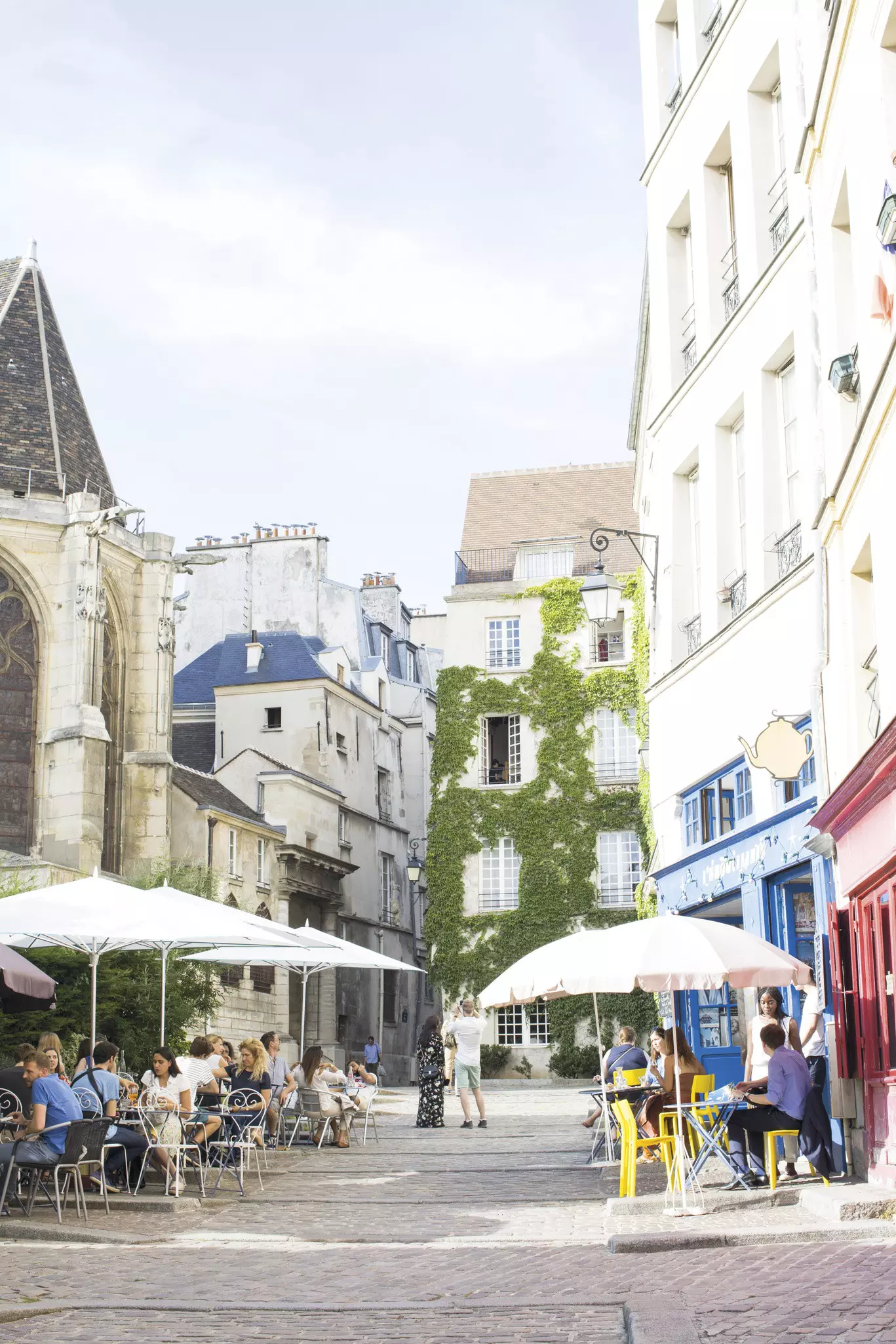People sit with drinks under large umbrellas at cafe terraces on a cobbled street near a church.
