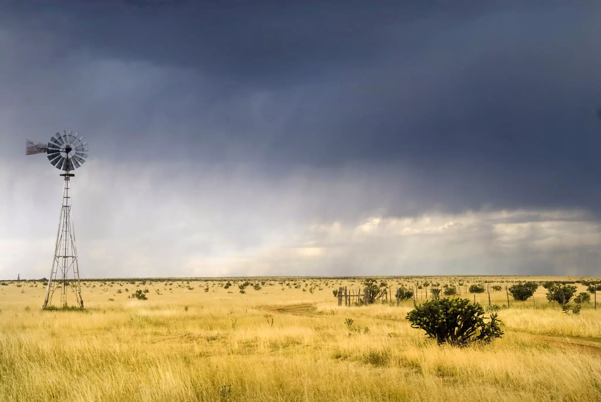 A windmill is pictured in a field of tall grass. Storm clouds loom overhead.