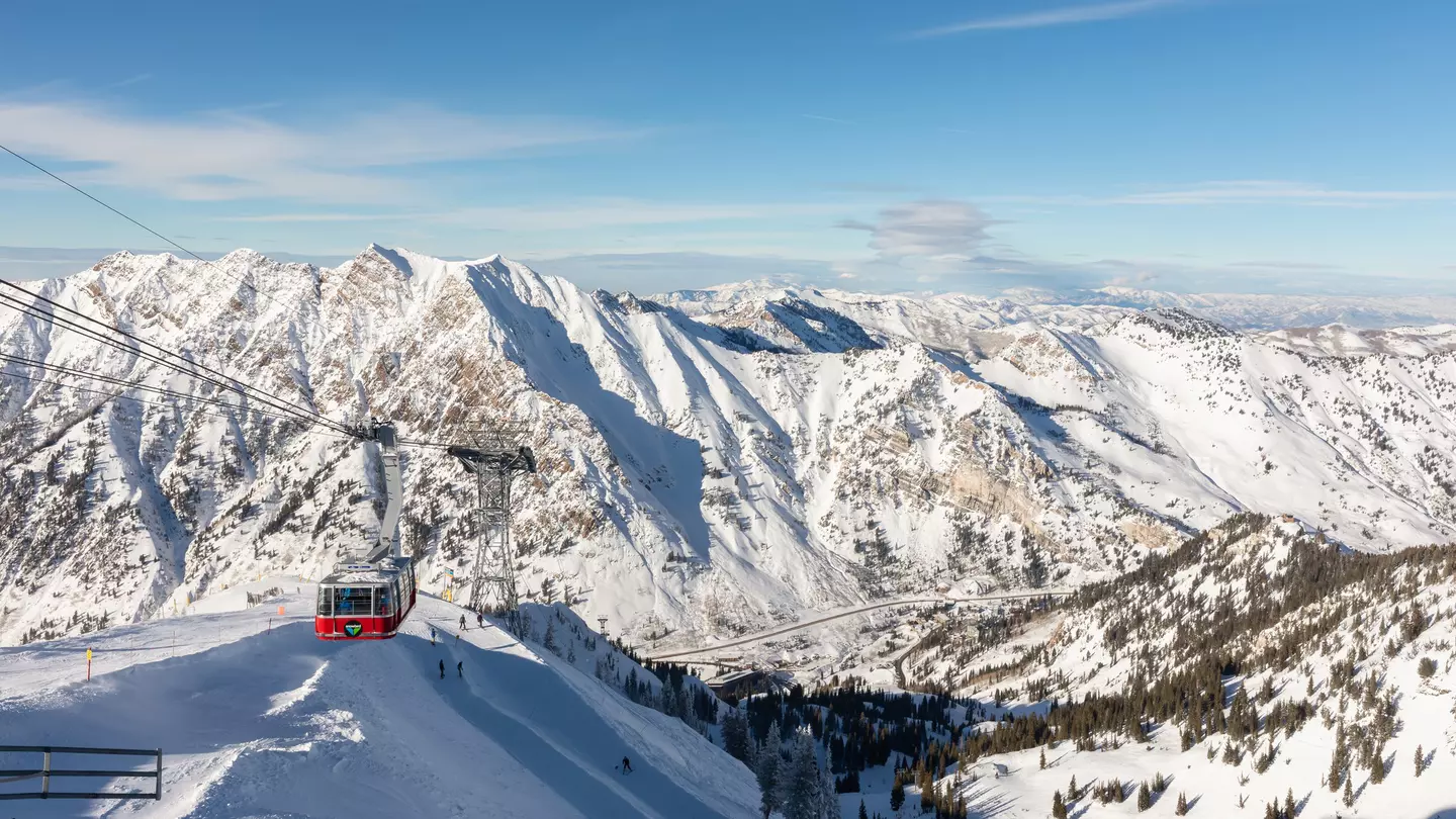 A wide view of an aerial tram ascending the snowy slope of a mountain, with other mountains in the distance.