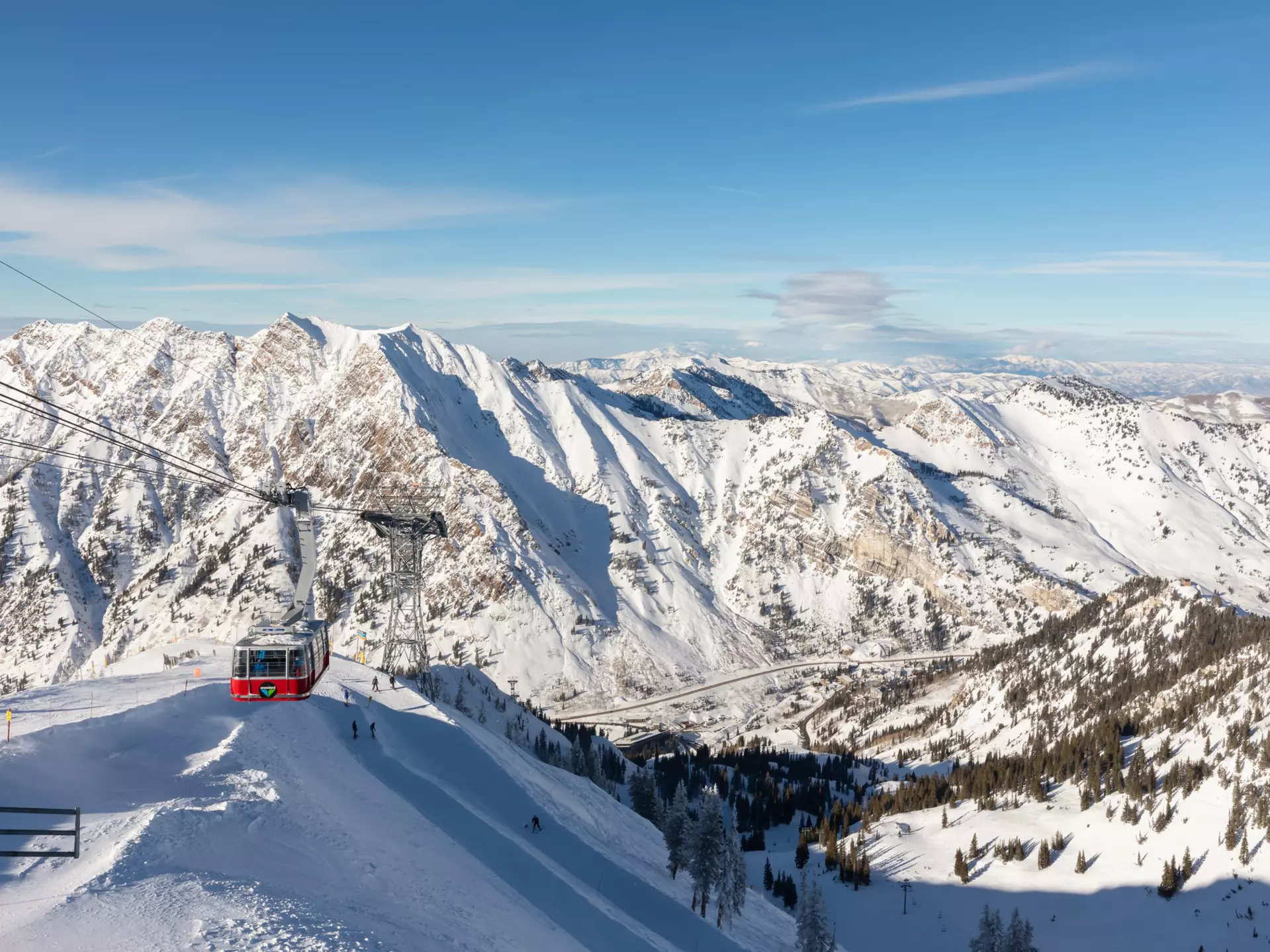 A wide view of an aerial tram ascending the snowy slope of a mountain, with other mountains in the distance.