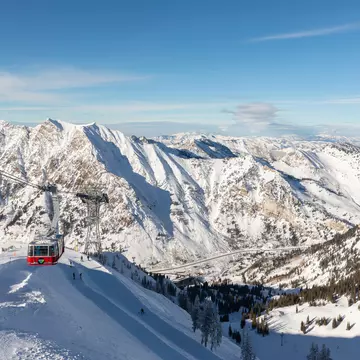 A wide view of an aerial tram ascending the snowy slope of a mountain, with other mountains in the distance.
