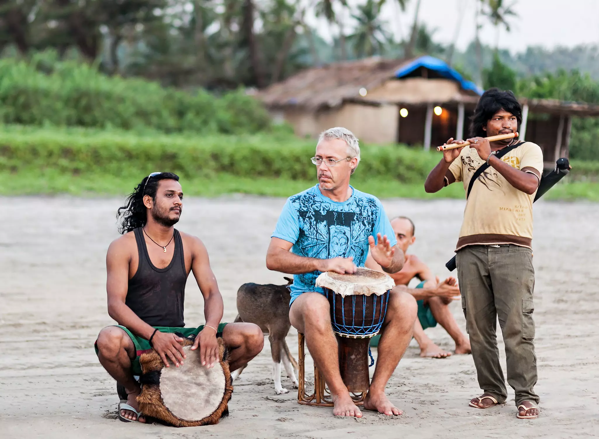 A group of men play drums on a beach. A structure and palm trees are visible in the background.
