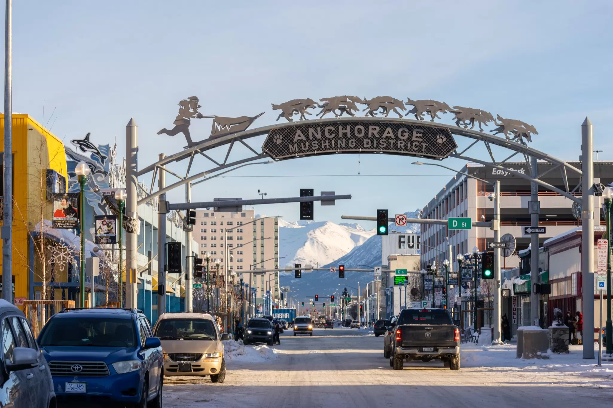 A gateway arch spans a wide street in the downtown of a city. Snowy mountains are seen in the distance.