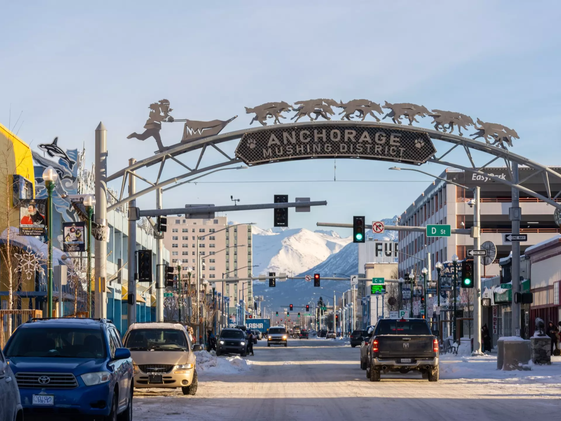 A gateway arch spans a wide street in the downtown of a city. Snowy mountains are seen in the distance.