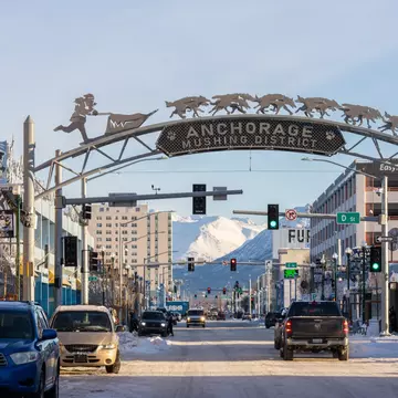 A gateway arch spans a wide street in the downtown of a city. Snowy mountains are seen in the distance.