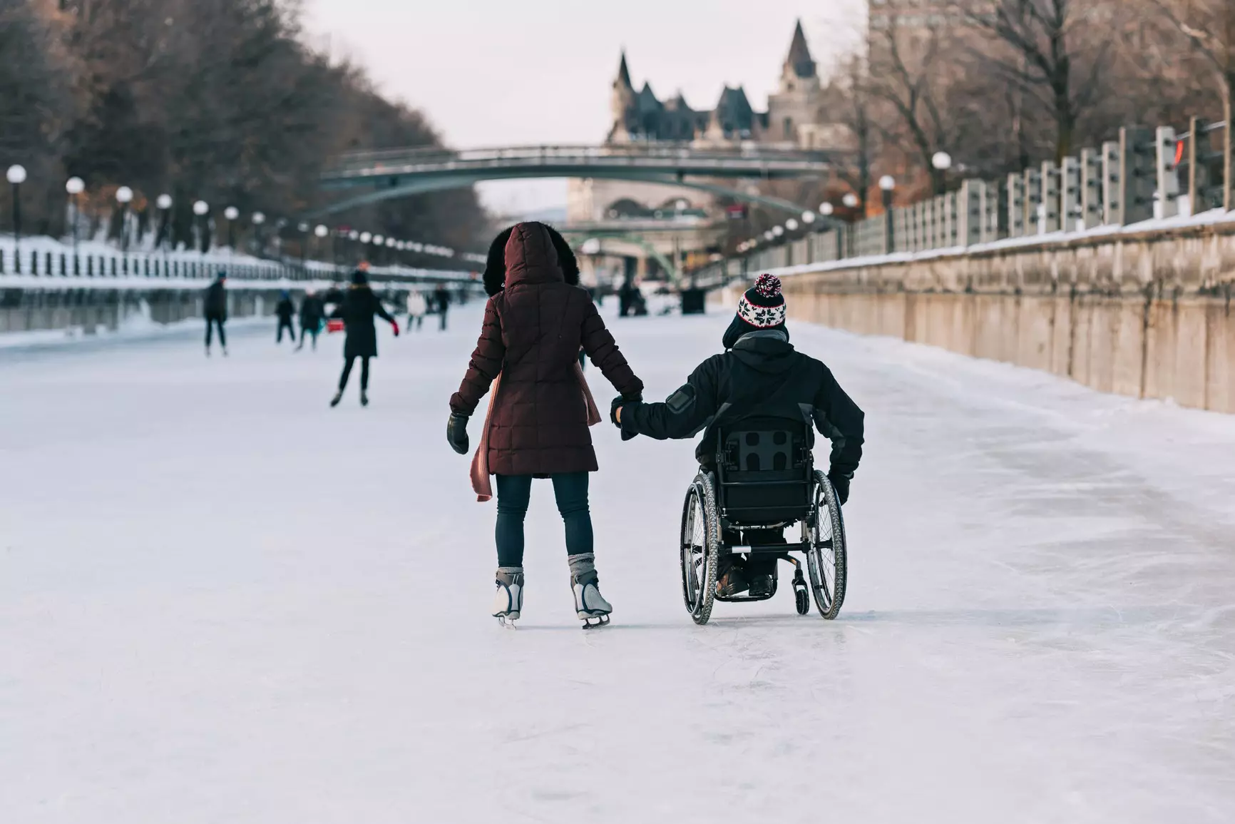 Man in wheelchair and woman skating on canal.