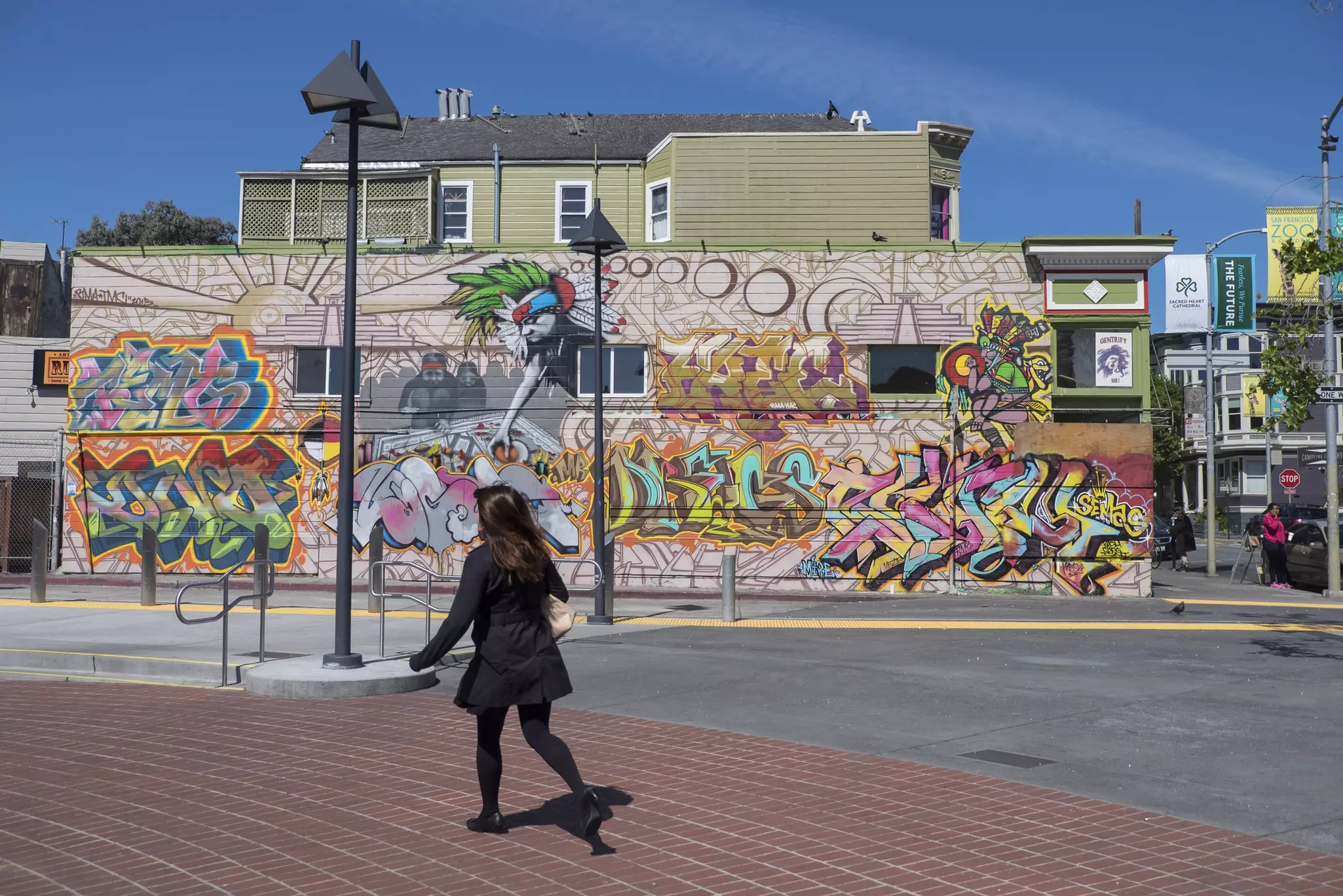 A woman in a black coat walks past a vibrant street mural on a sunny day.