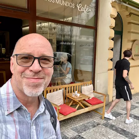 A man smiling in front of a glass-front cafe that has a wood bench with cushions in front