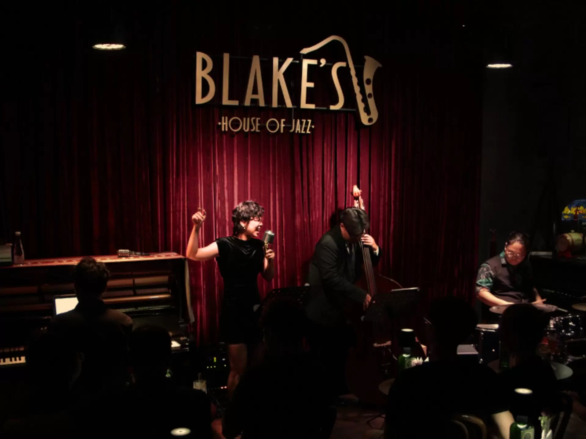 A darkened room with performers in black onstage in front of a red curtain with the name "Blake's House of Jazz" and a saxophone on it in gold