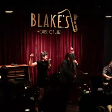 A darkened room with performers in black onstage in front of a red curtain with the name "Blake's House of Jazz" and a saxophone on it in gold