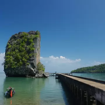 Longtail boat on clear green water next to a long concrete pier and a tall limestone rock covered in trees, under a blue sky.