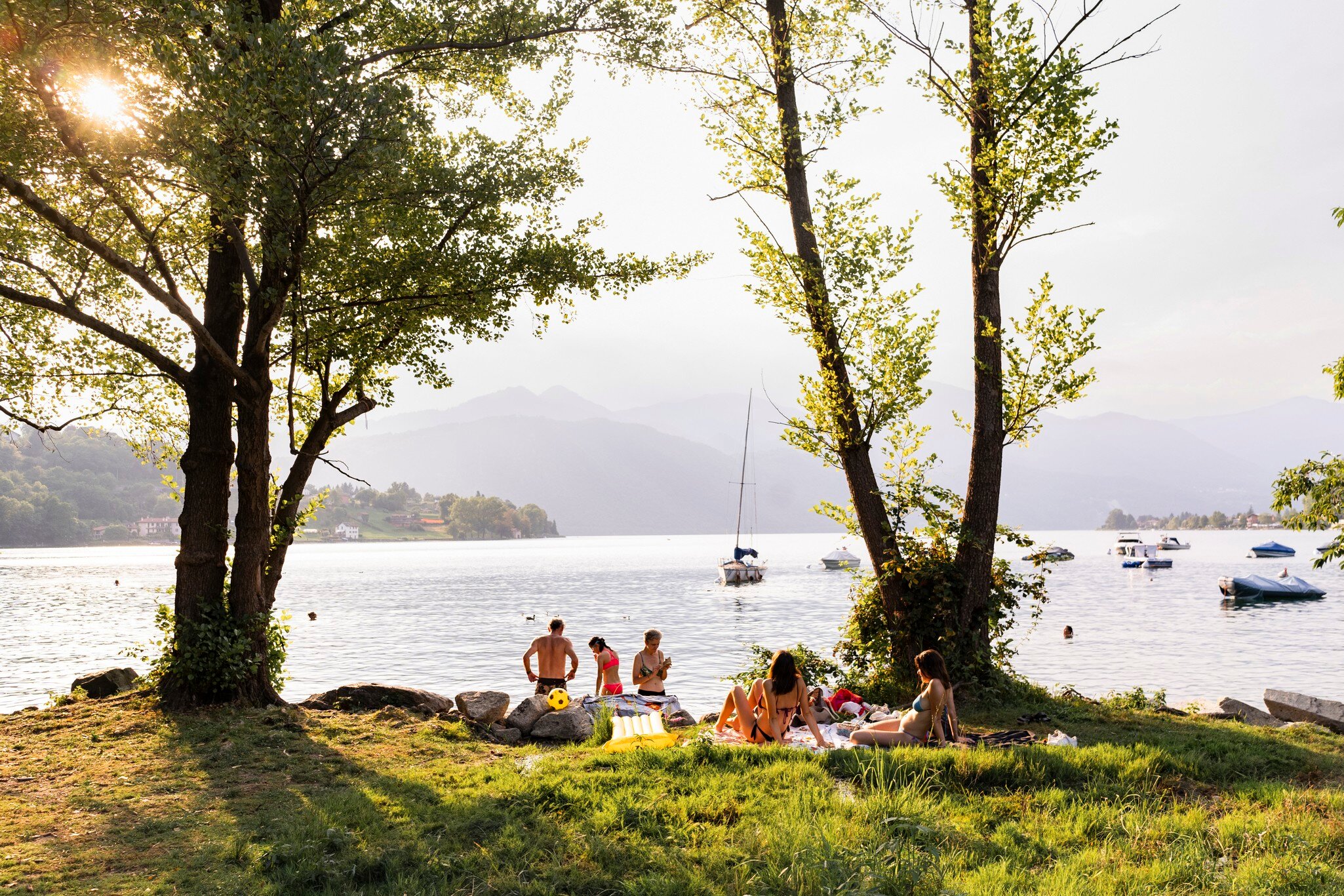 Friends relax on grass under trees on the shore of Lake Orta, Piedmont, Italy, with boats and hazy hills behind