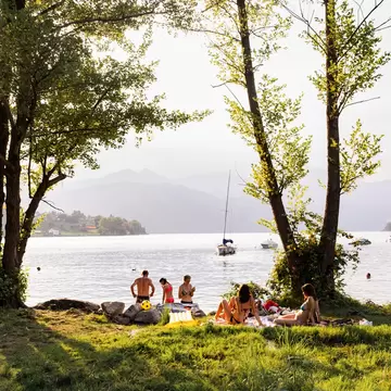 Friends relax on grass under trees on the shore of Lake Orta, Piedmont, Italy, with boats and hazy hills behind