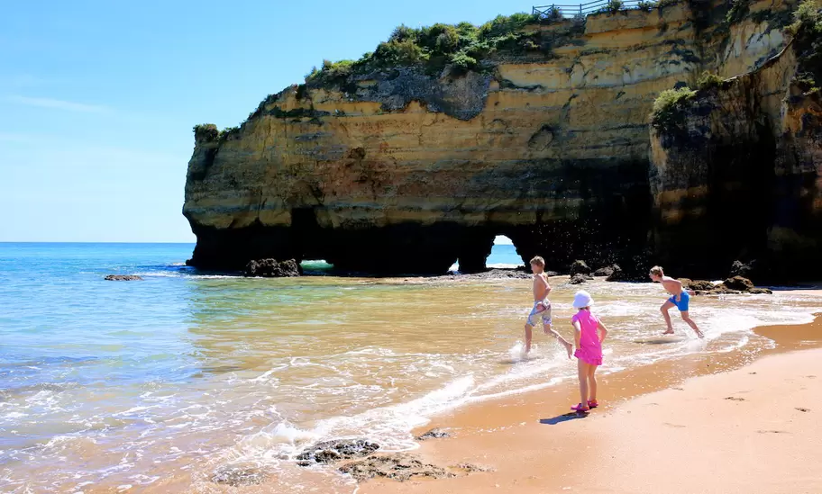 Golden sandstone cliff with sea arches on a Lagos beach in the Algarve, Portugal, as three children run into the shallows