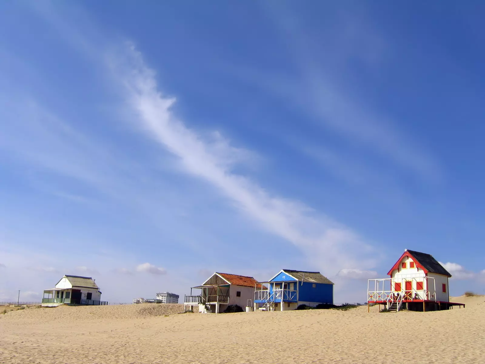 Small, colorfully painted cabins sit on a wide beach under a blue sky.