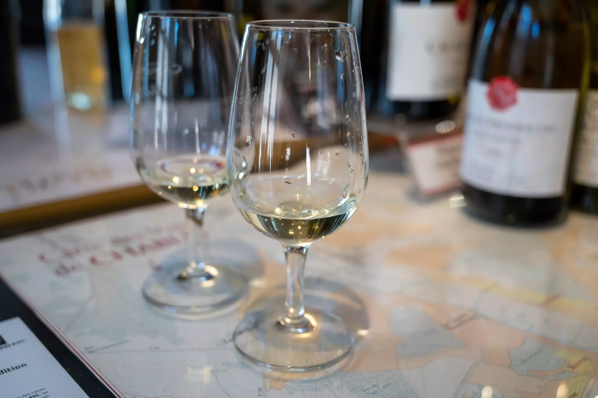 Two wine glasses on a table each holding a small tasting of pale white wine.