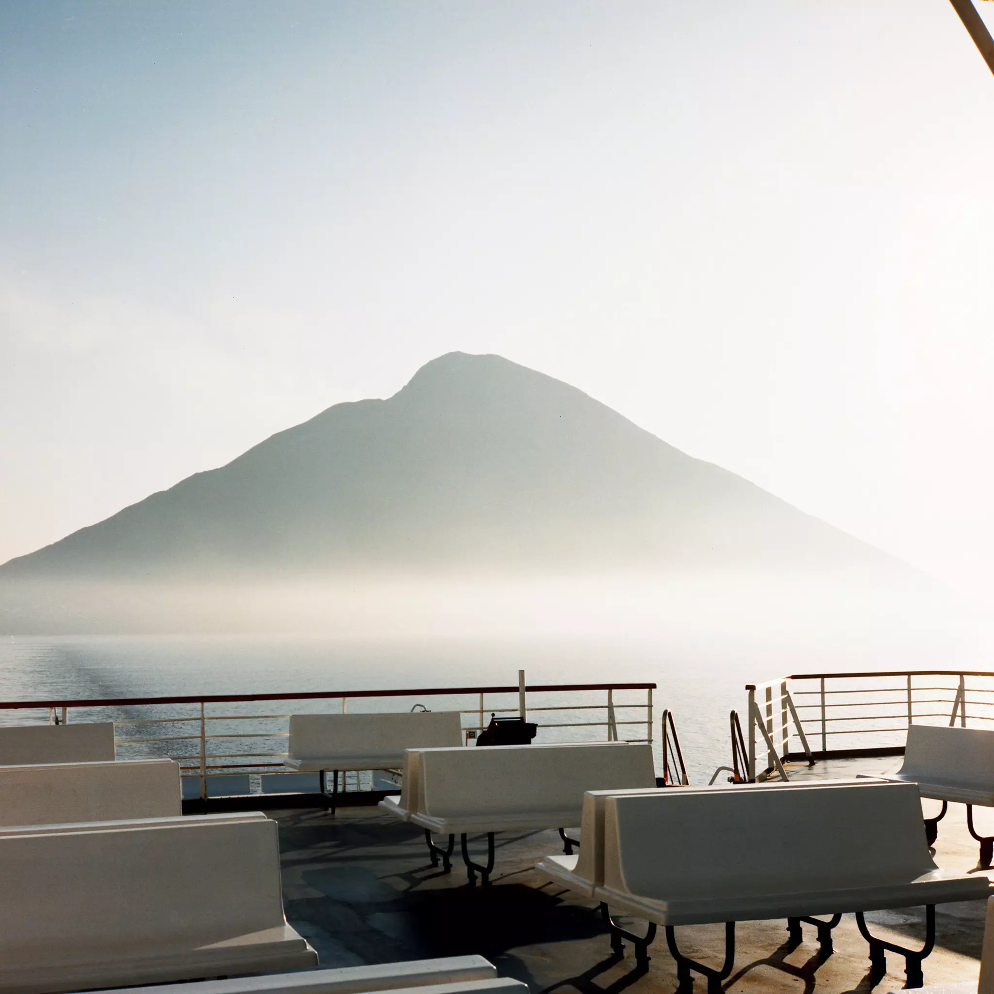 A volcano shrouded in mist is seem from the empty deck of a ferry on the water.