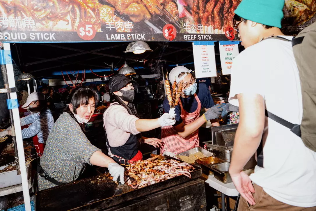 Three people work at a food stand. One hands skewers of food to a customer.