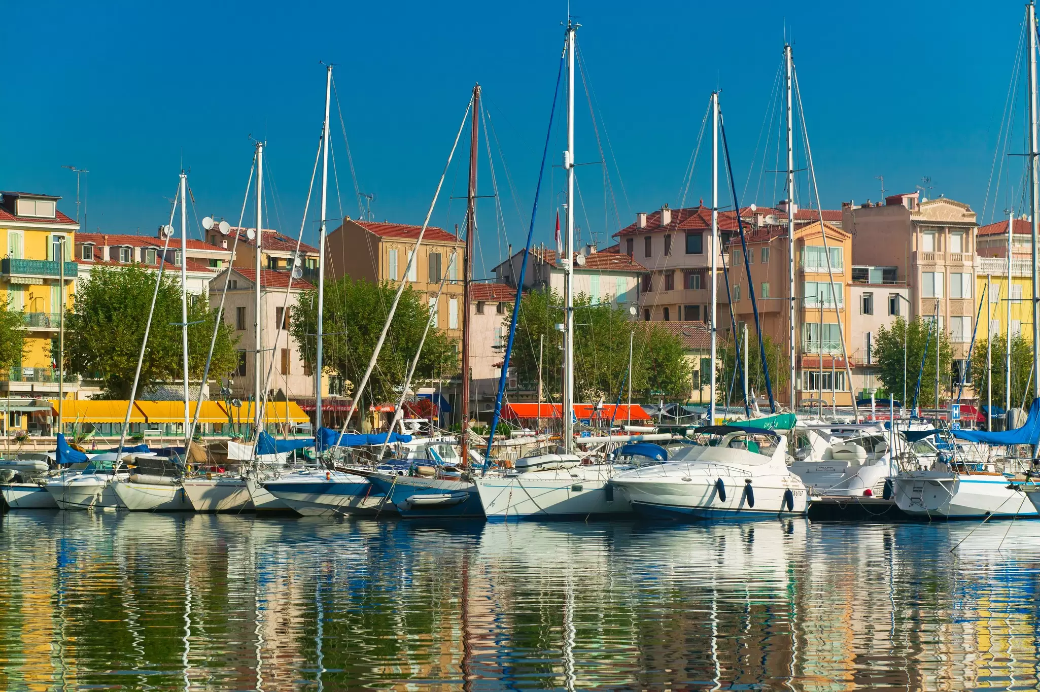 Boats in a harbor are reflected in the water; buildings on the shore are in bright pastel tones under blue sky.