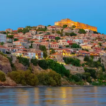 Stone houses with tiled roofs climb a hillside to a floodlit hilltop castle at dusk, above calm water.