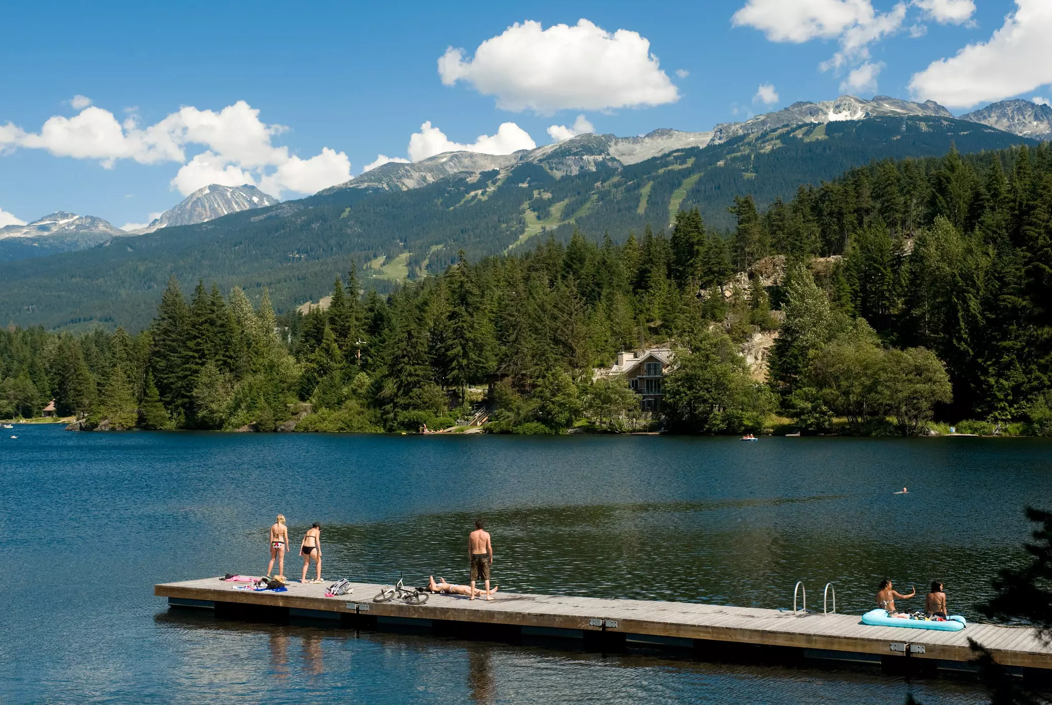 People in swimsuits at the end of a wooden pier over a lake in Canada; mountains are on the opposite shore.