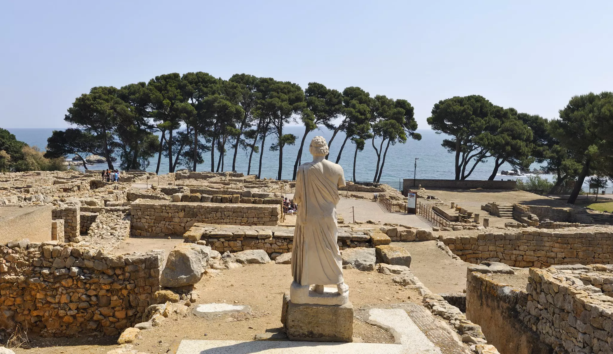 A large marble statue of a person wearing a toga stands among ancient ruins at an archaeological site facing a line of trees and the sea.