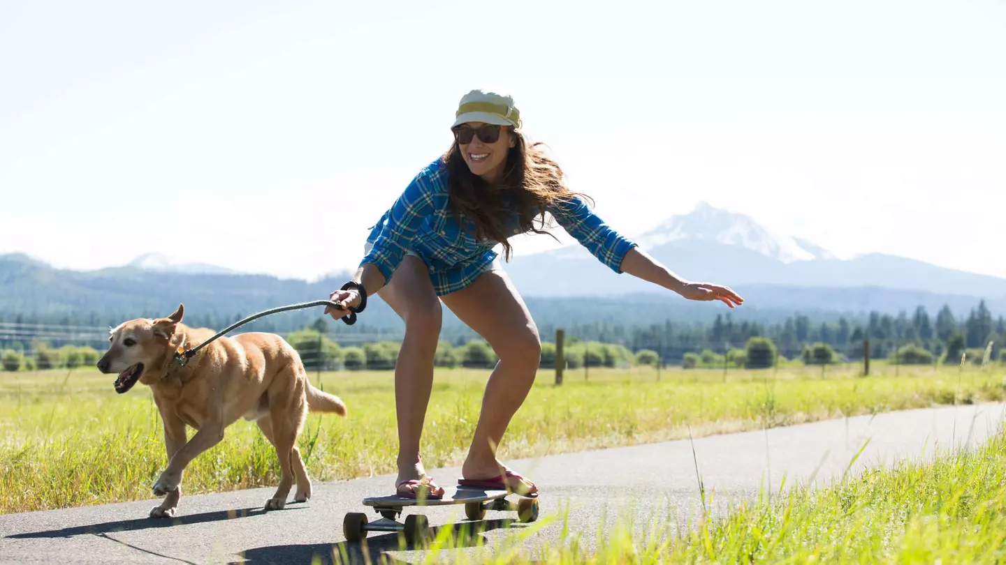A woman skateboarding with her dog pulling her and a big mountain visible behind them