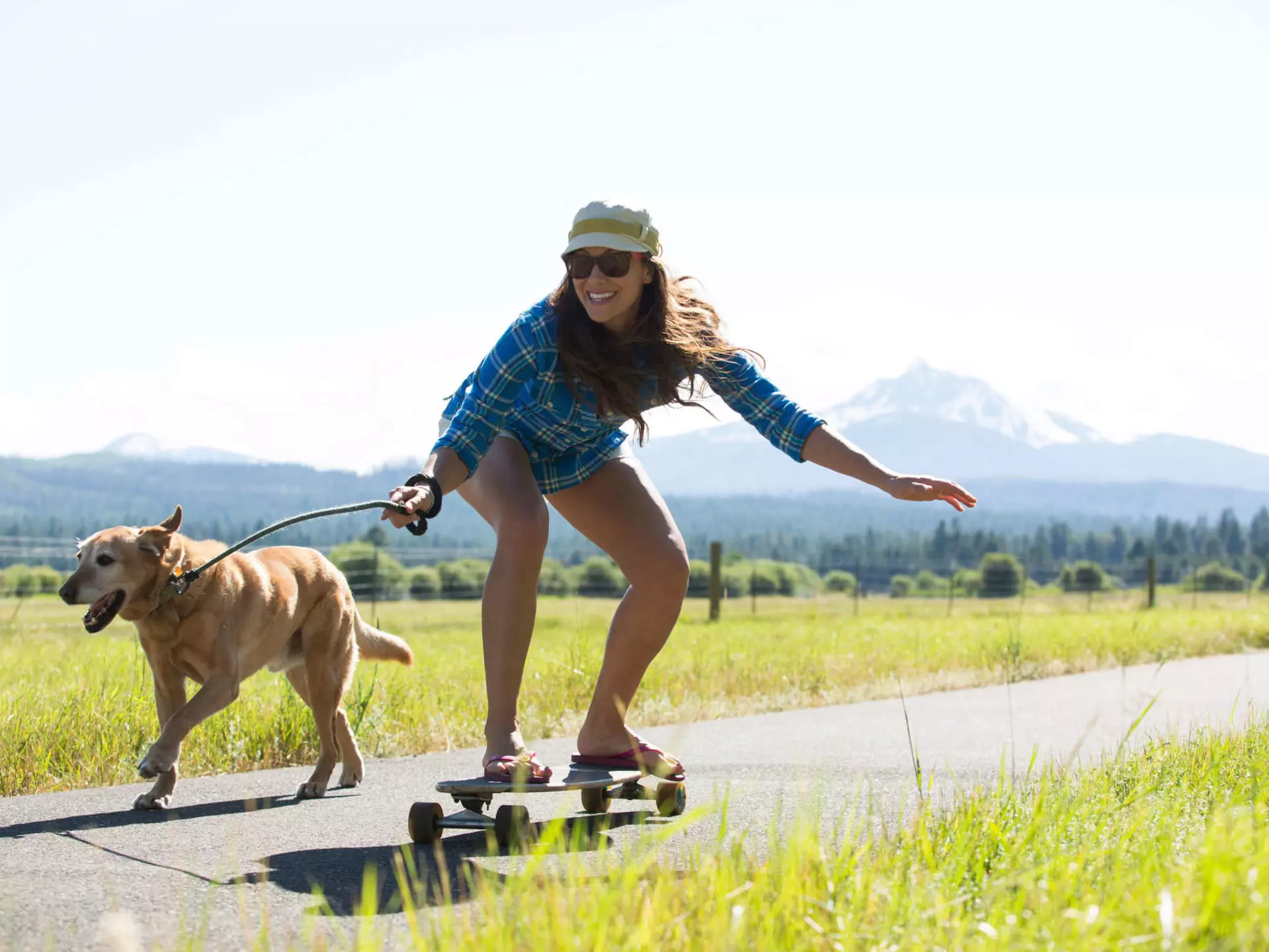 A woman skateboarding with her dog pulling her and a big mountain visible behind them