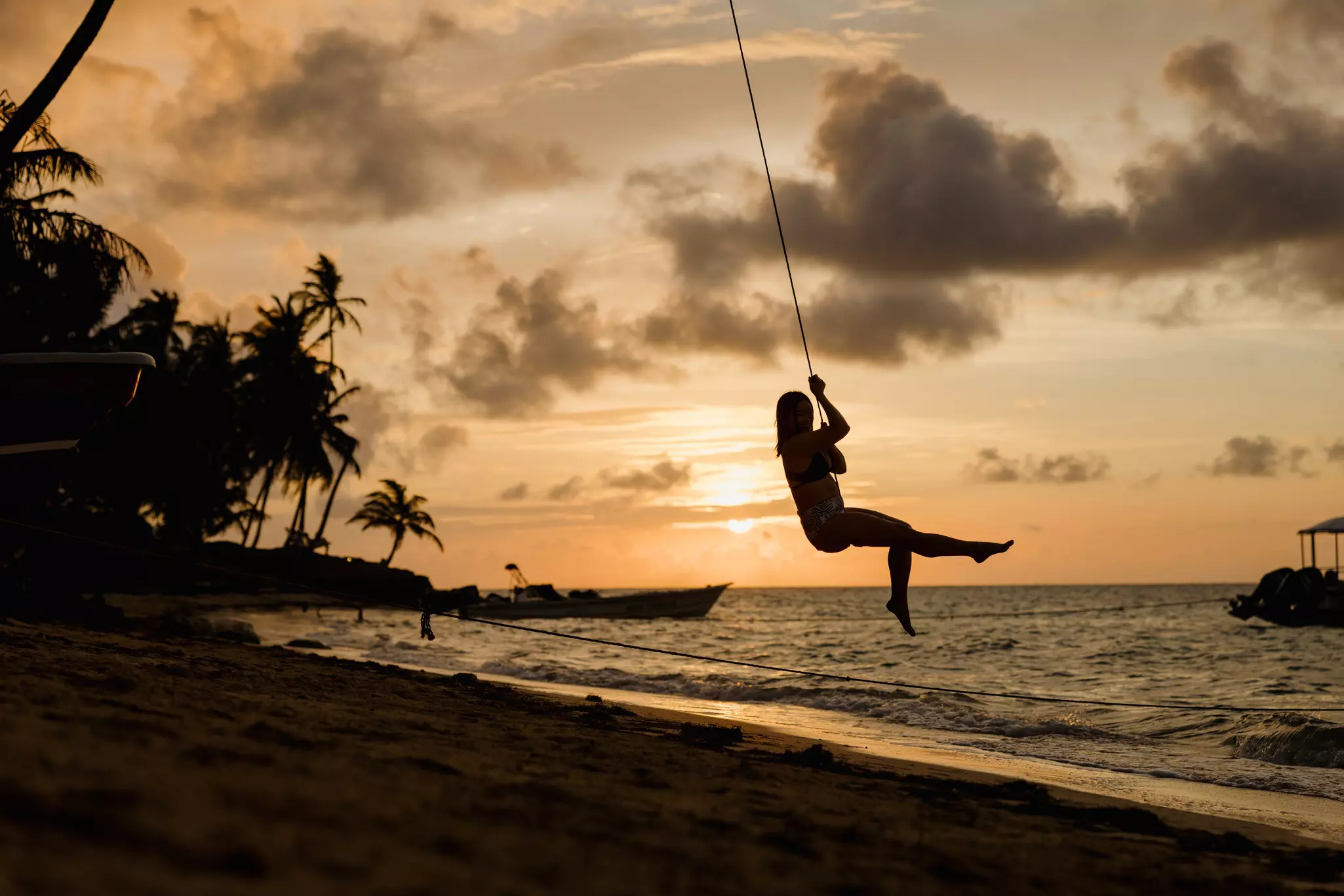 Woman on a swing tied to a palm tree at sunset on a beach.