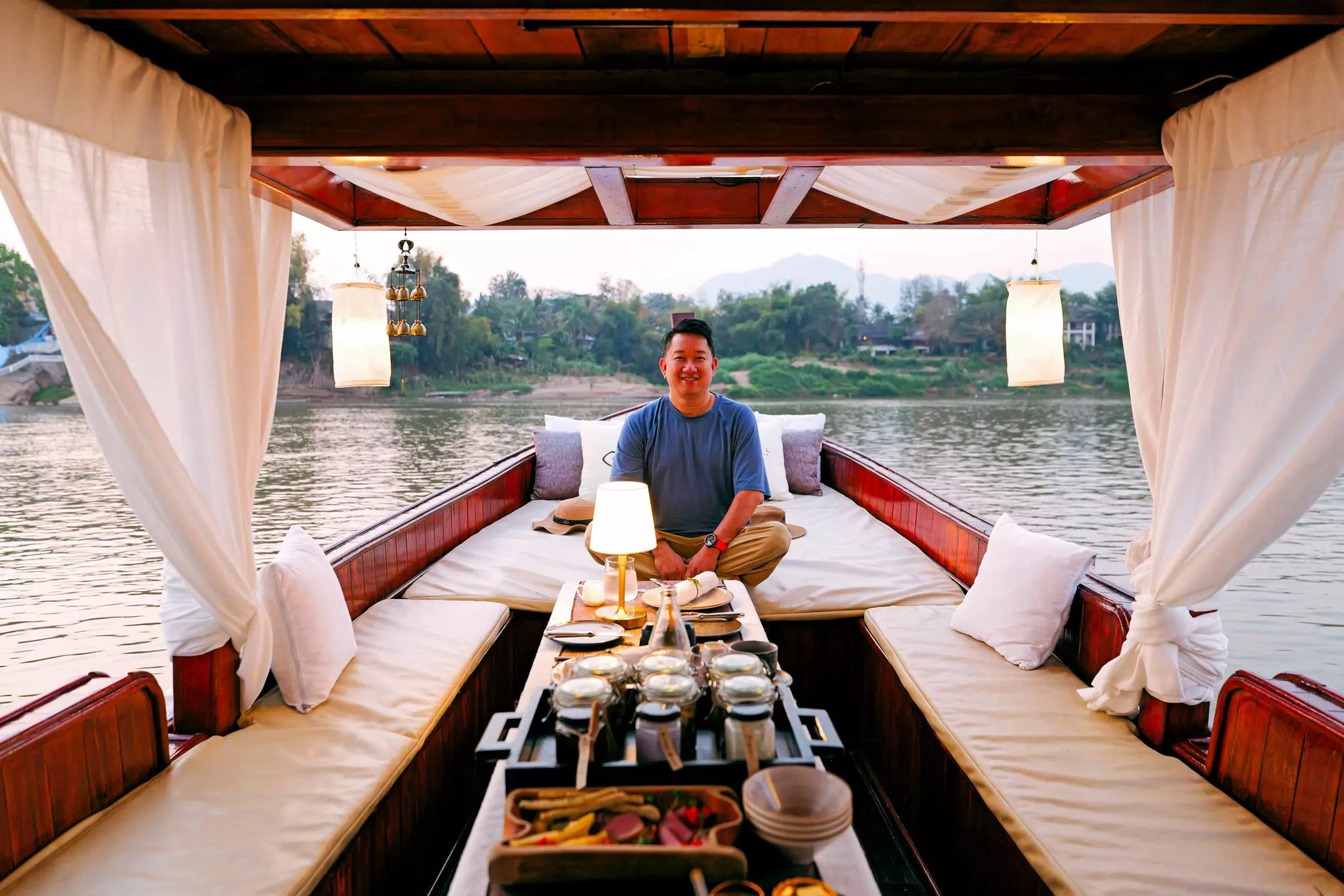 A man sits on comfy lounge seats on a wooden boat that's floating on a river. Breakfast is laid on the table