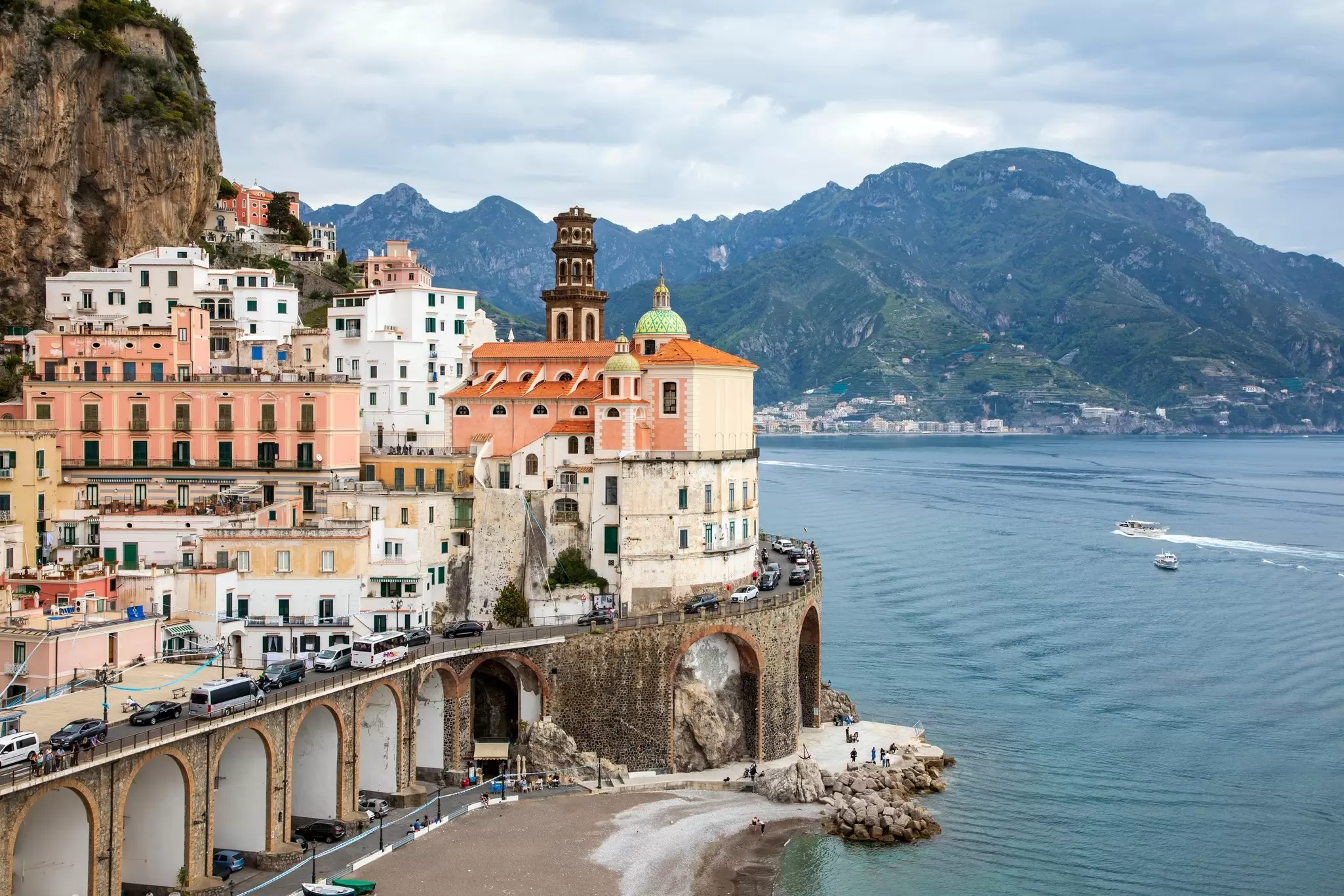 A cluster of buildings in a coastal village. Vehicles travel a winding road around the buildings, Boats are visible on the water just off the coast, and a verdant mountain rises in the background.