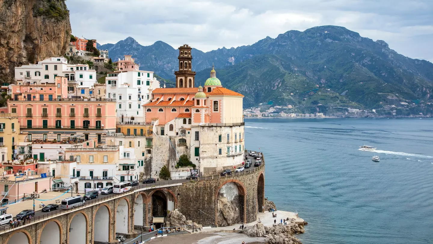 A cluster of buildings in a coastal village. Vehicles travel a winding road around the buildings, Boats are visible on the water just off the coast, and a verdant mountain rises in the background.