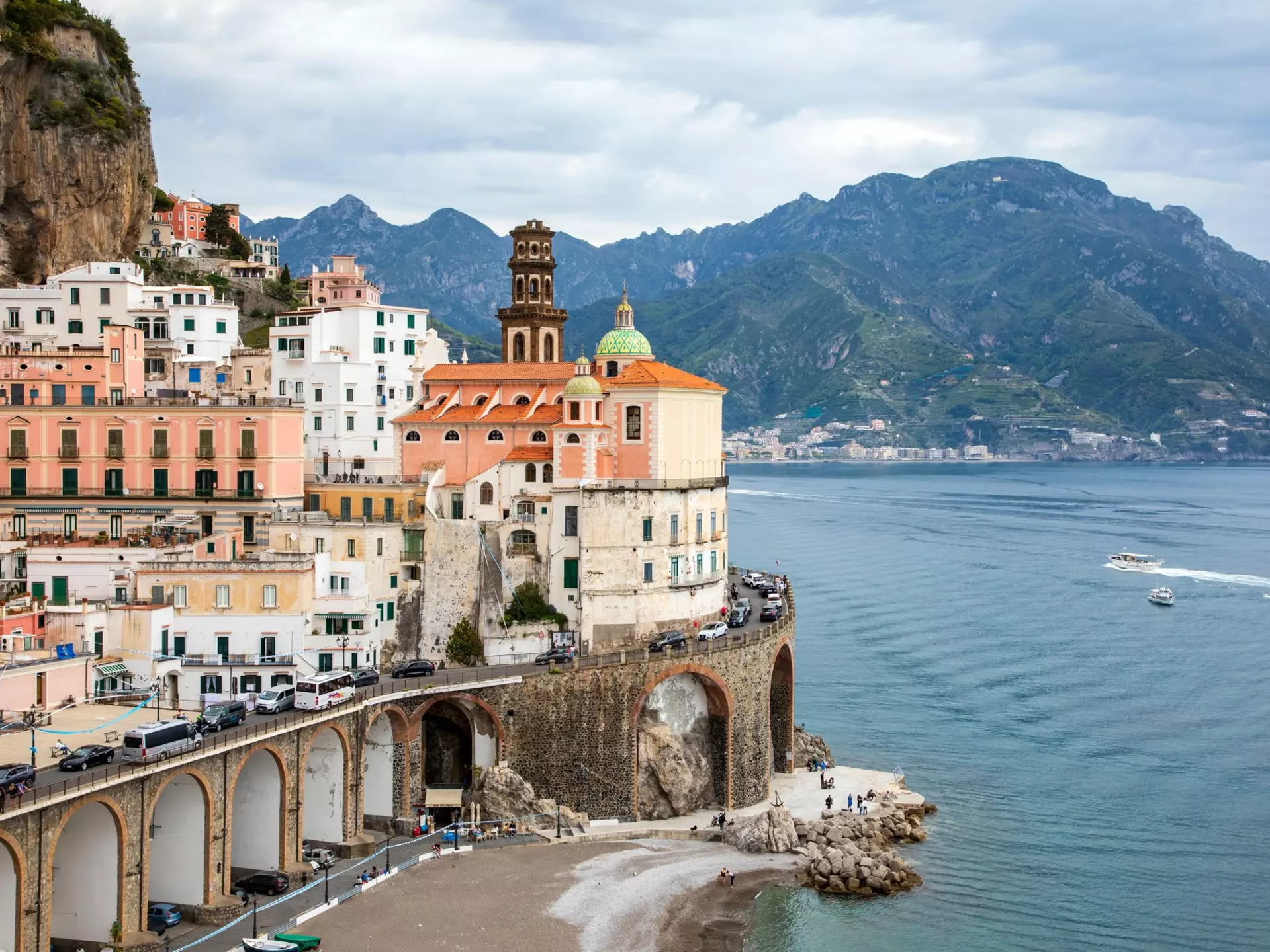 A cluster of buildings in a coastal village. Vehicles travel a winding road around the buildings, Boats are visible on the water just off the coast, and a verdant mountain rises in the background.