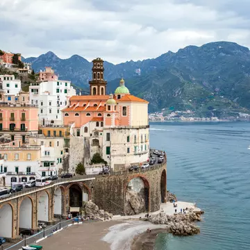 A cluster of buildings in a coastal village. Vehicles travel a winding road around the buildings, Boats are visible on the water just off the coast, and a verdant mountain rises in the background.